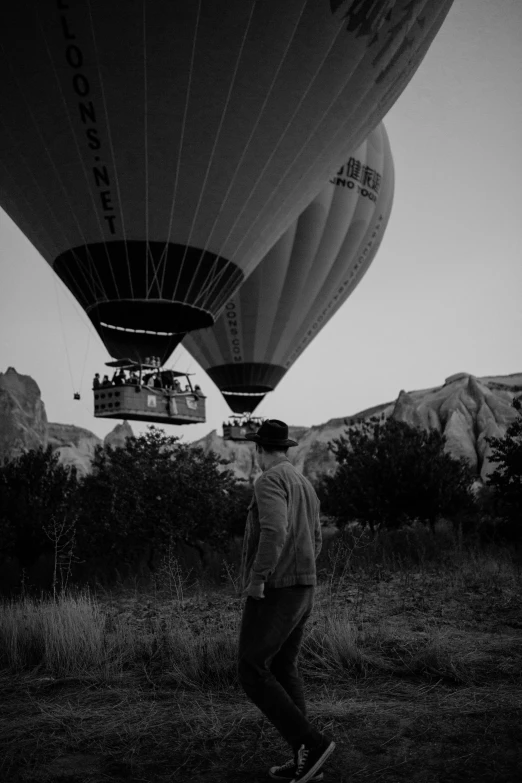 a man stands on the grass with some  air balloons flying in the background