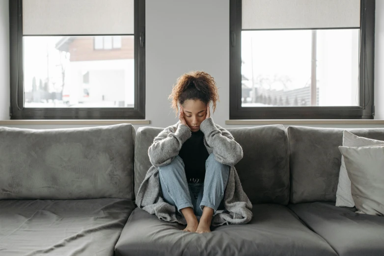 a woman sits on a couch in front of windows with her hands to her head