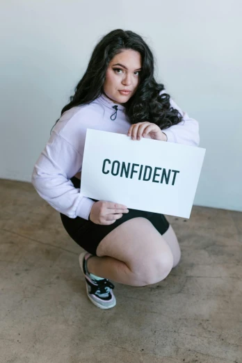 a woman squatting in front of a white sign that says confident
