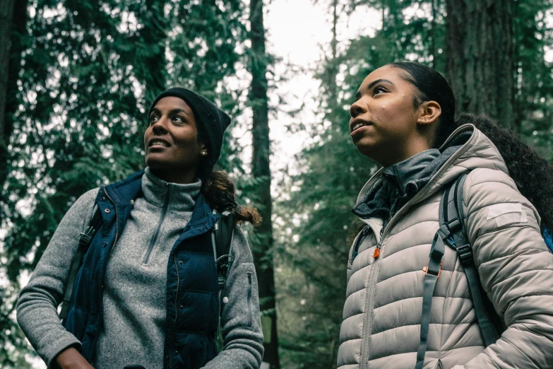 two women stand in the woods and look to the side