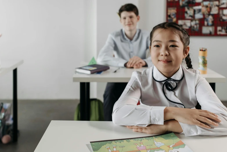 two children sit at a table, one is wearing a white shirt and the other a grey tie