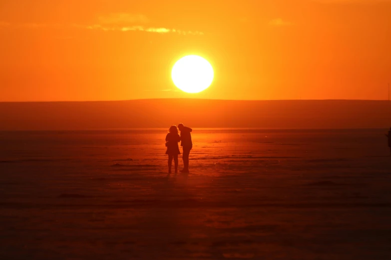 the people are standing on the beach while the sun sets