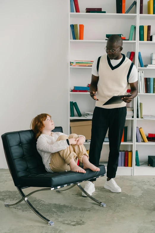 two women sit on an office chair, with a book shelf behind them