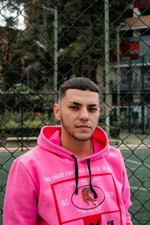 a young man standing in front of a fence wearing a pink hooded sweatshirt