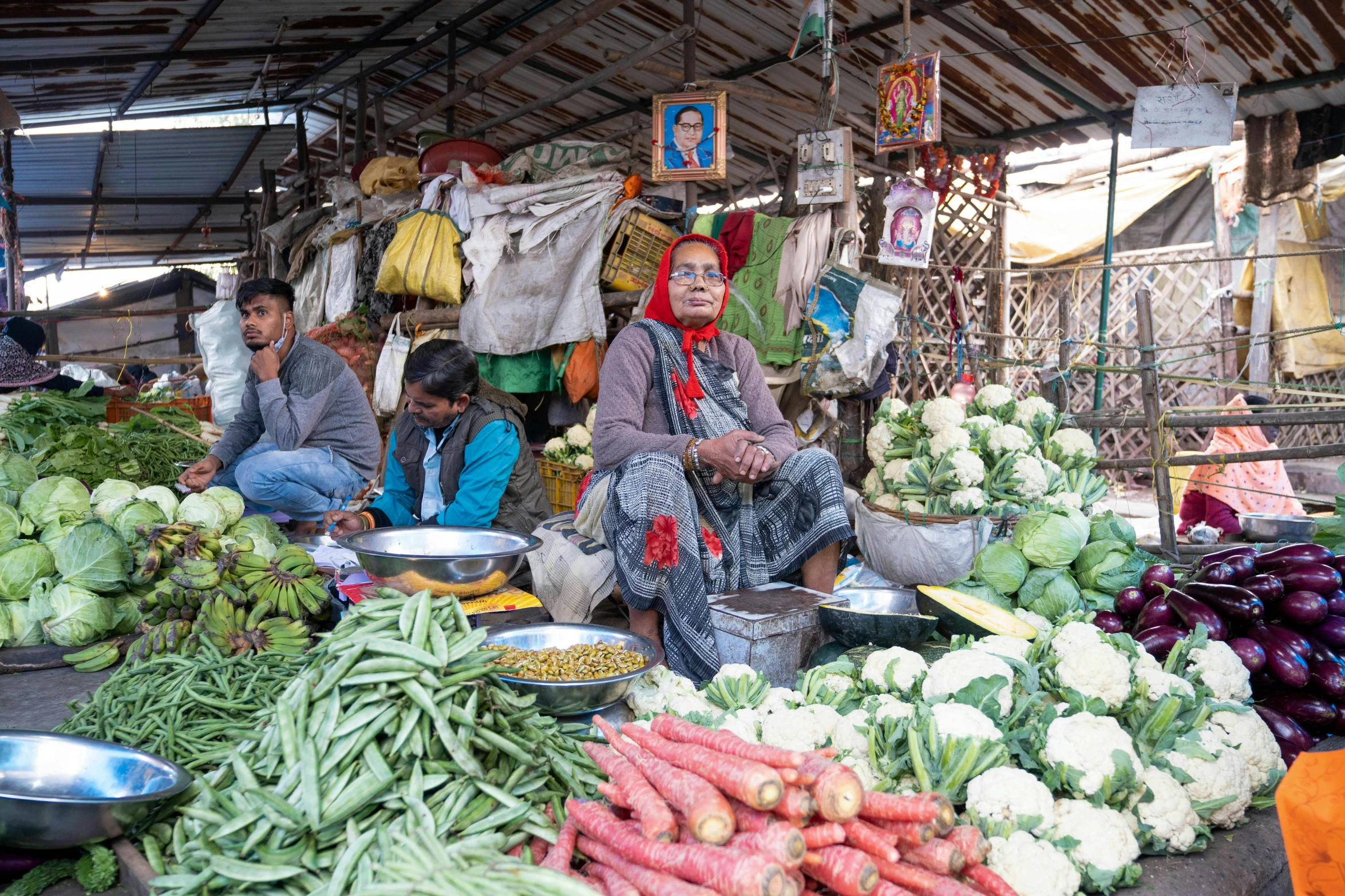 a woman sits at a vegetable market while other people are gathered around