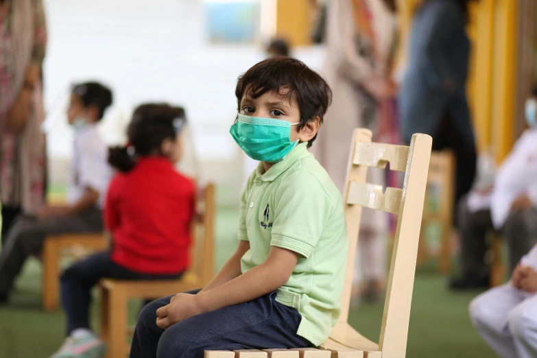 little boy wearing a green shirt and face mask on a wooden chair