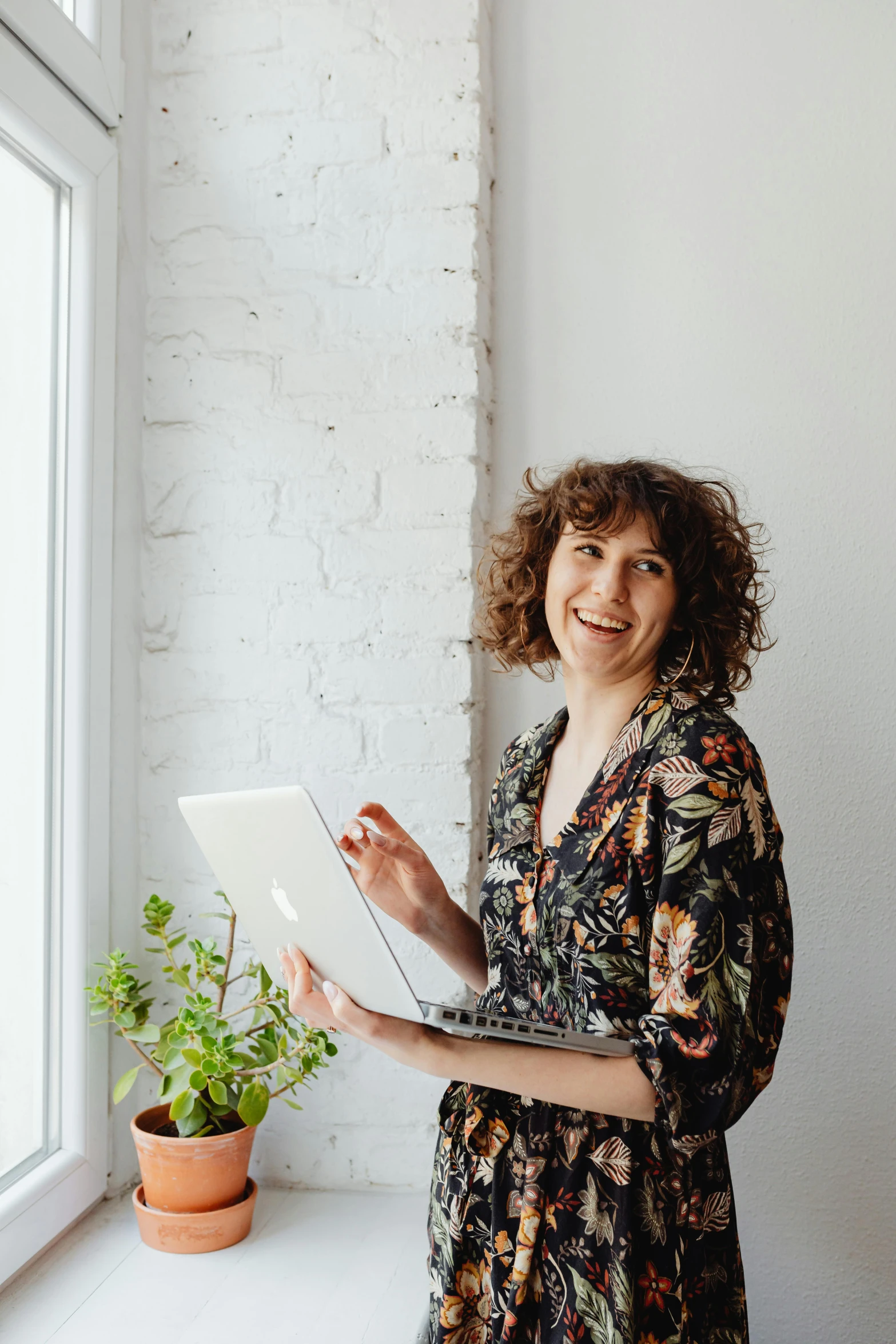 a woman stands next to a window holding a laptop