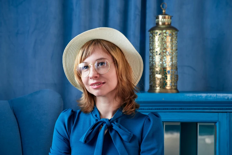 a woman wearing a blue dress sitting in a chair wearing a white hat and glasses