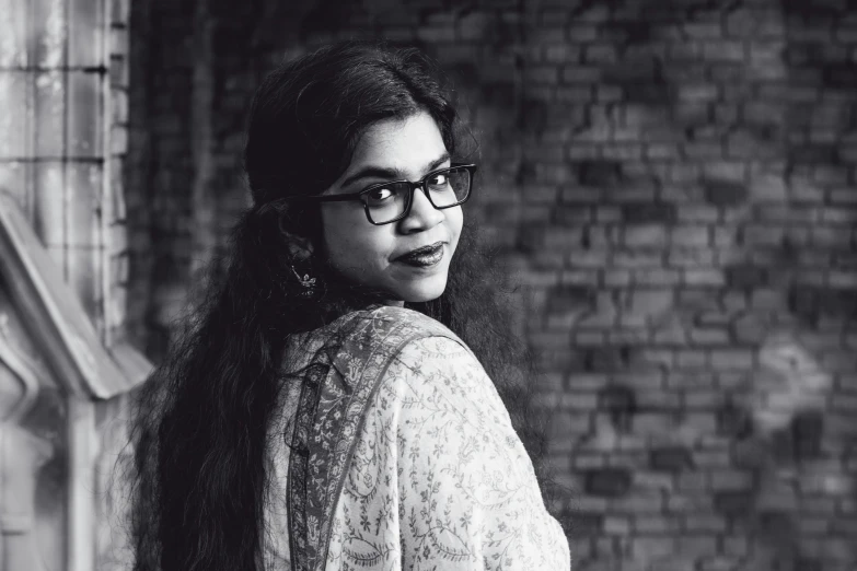 a woman with glasses and sweater standing in front of brick wall