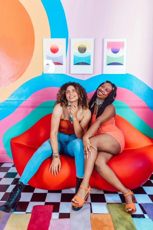 two women sitting on a rainbow - painted couch in an art - filled room