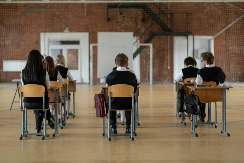 three students are sitting down at desks in a gymnasium