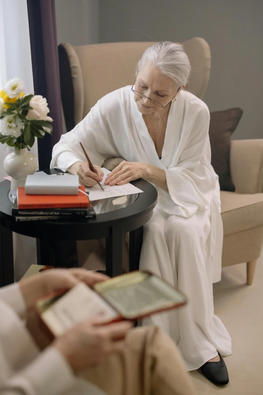 an elderly woman sitting at a table writing a book