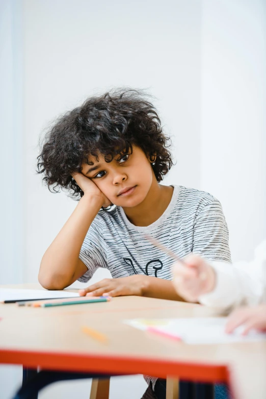 a young person sits at a table, while someone writes on a piece of paper