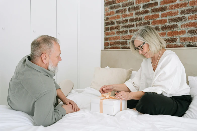 an old man and older woman sitting on a bed giving each other gifts