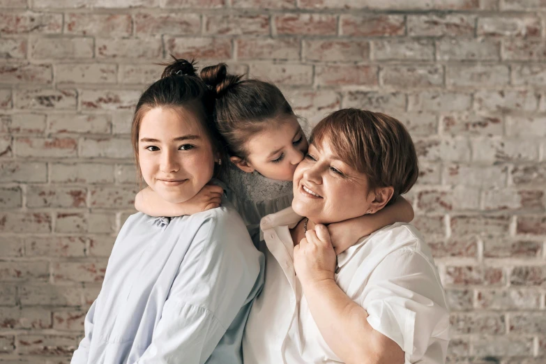 a mother kisses her daughters cheek as they lean against a brick wall