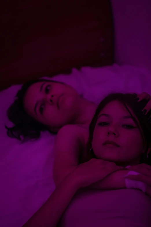 two women laying in a bed, one woman is dark purple lighting