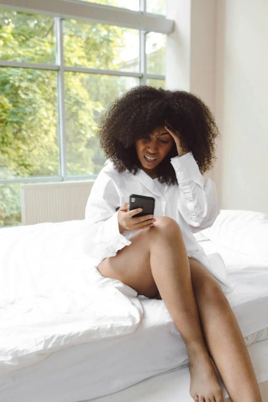 a woman in white shirt sitting on a bed and holding a cell phone