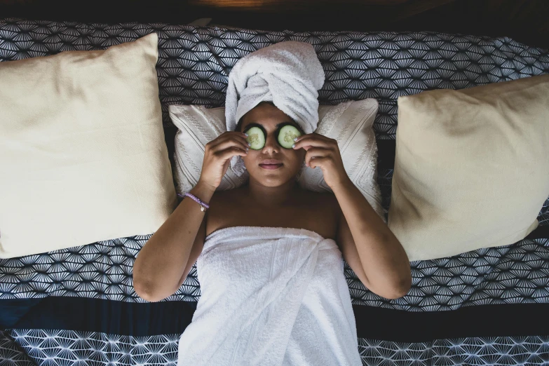 the girl is lying on the bed and cucumbers have a towel on her head