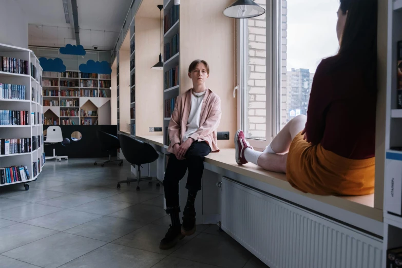 a man sits on a desk while another man stands at the window