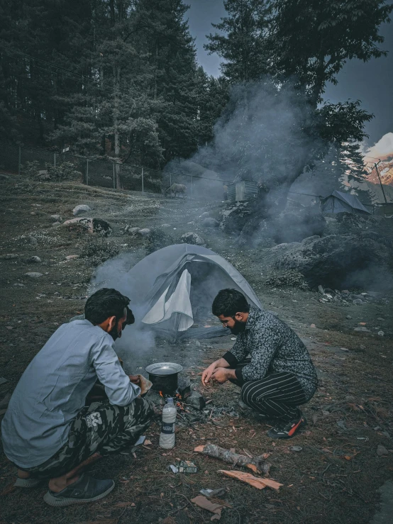 two men sitting on the ground in front of a campfire with a stove