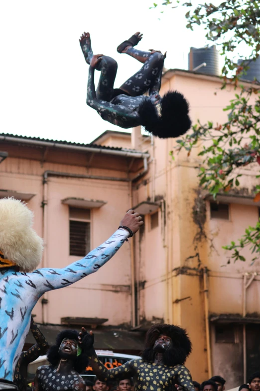 a person on stilts, with painted clothes and headdress is performing a trick on the same pole