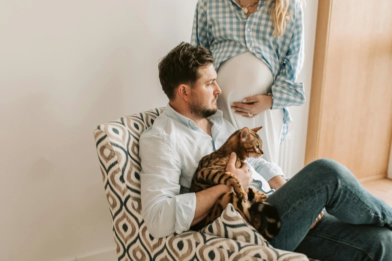 the young man is sitting next to his pregnant wife and holding their cat