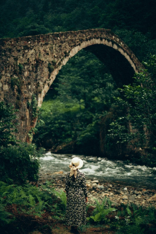 a woman in floral dress standing by stream under an arched stone bridge