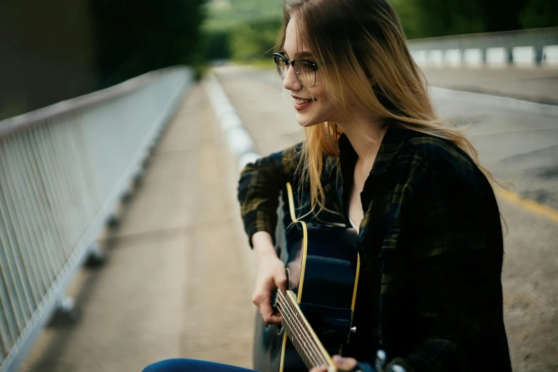 a girl with glasses holding a guitar sitting on the side of the road