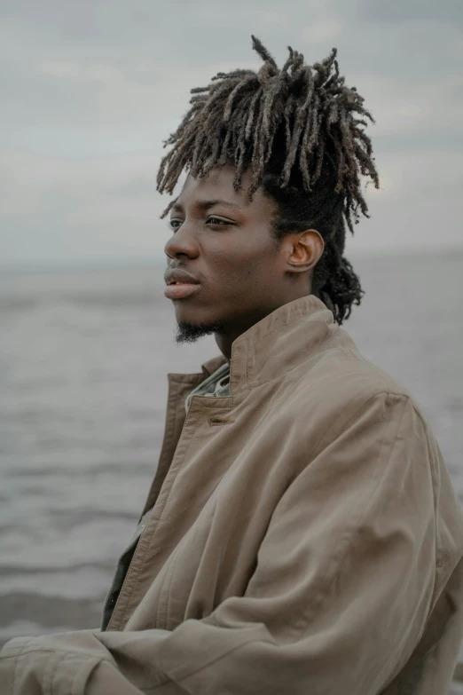 man in brown jacket with dreadlocks sitting next to water