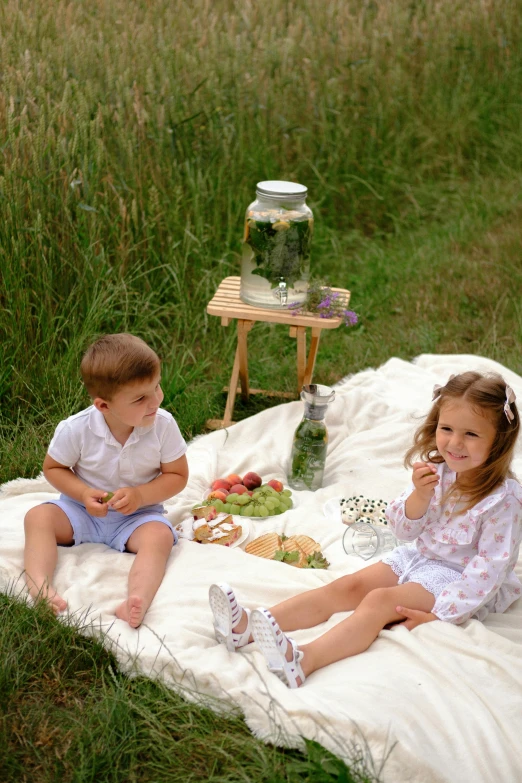 two children sit on a blanket eating food