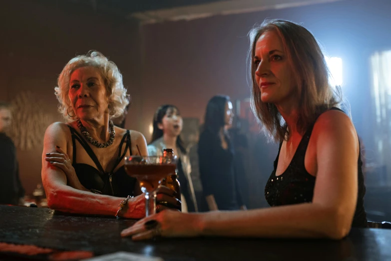 two women in dress clothes sit at a bar with other people