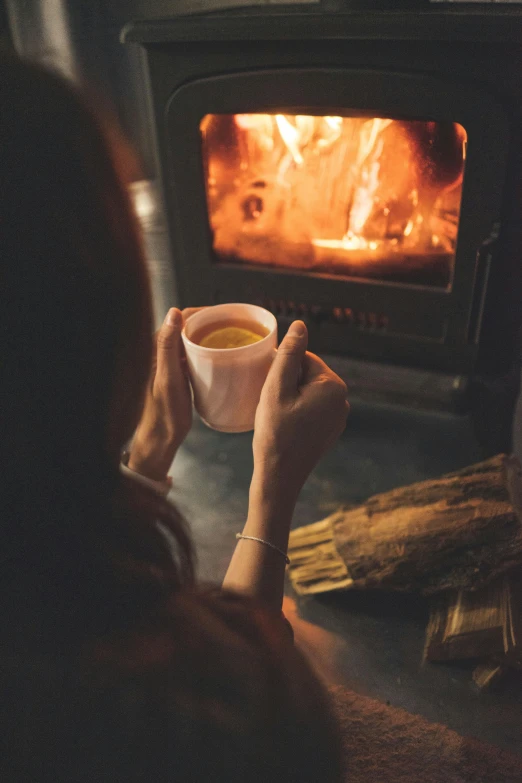 a woman holding up a cup of tea and sitting in front of an open wood stove