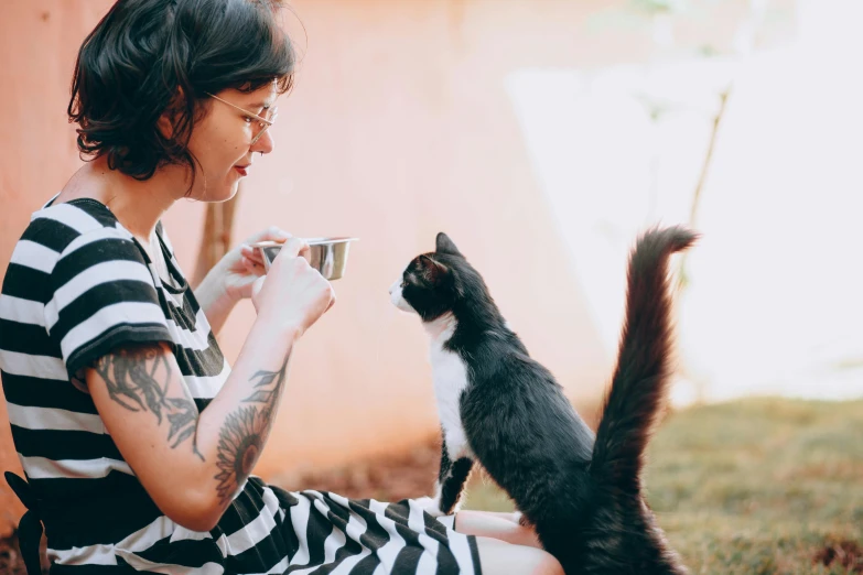 the woman with black and white striped dress is drinking coffee from a coffee cup with her cat, looking on