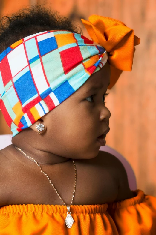 a young baby girl wearing an orange top and headband with a big piece of jewellery on it's left side