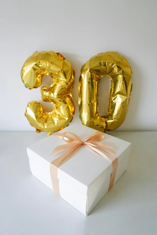 a white gift box with golden foil balloons and a bow
