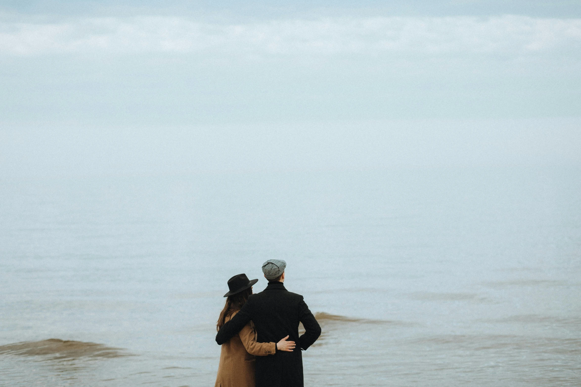 couple in long coats standing on the beach next to water