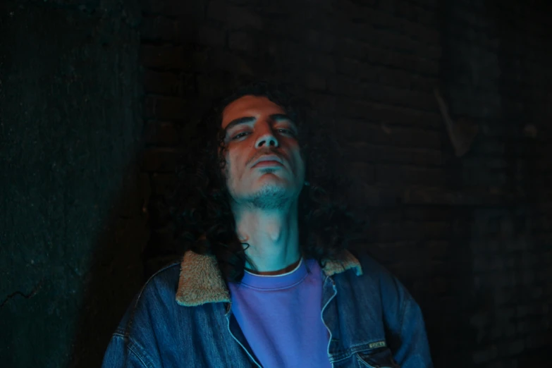 a man with long hair and blue shirt standing next to a brick wall