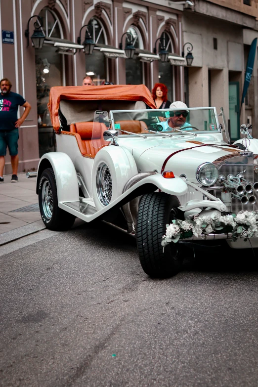 the silver car is going down the street and people are waiting in line