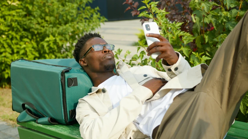 a man relaxing on a green bench and taking a selfie