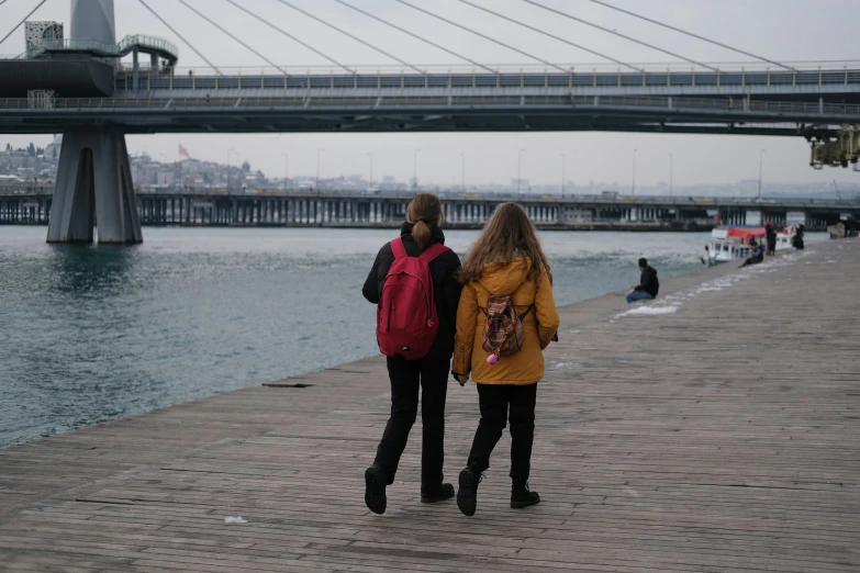 two people walking in front of the water under an overpass