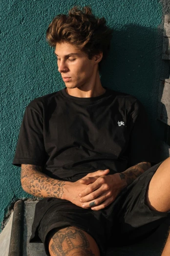 a male wearing tattoos sits on the ground next to a green wall