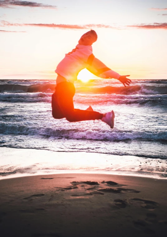 a woman jumps from the water onto a sandy beach as the sun sets