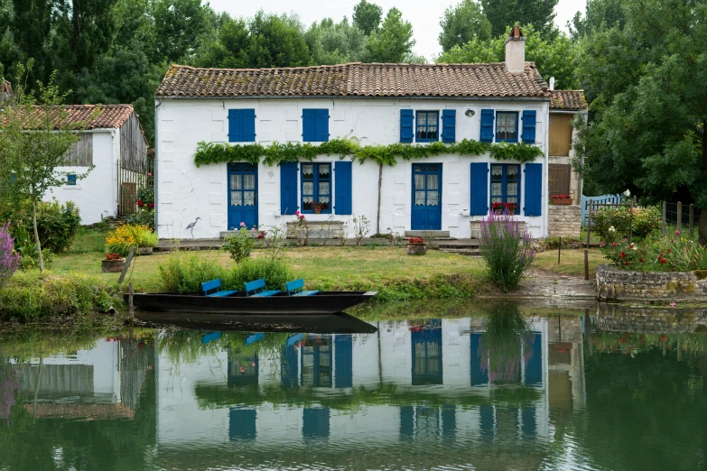 a boat sits on a river near a white house with blue shutters