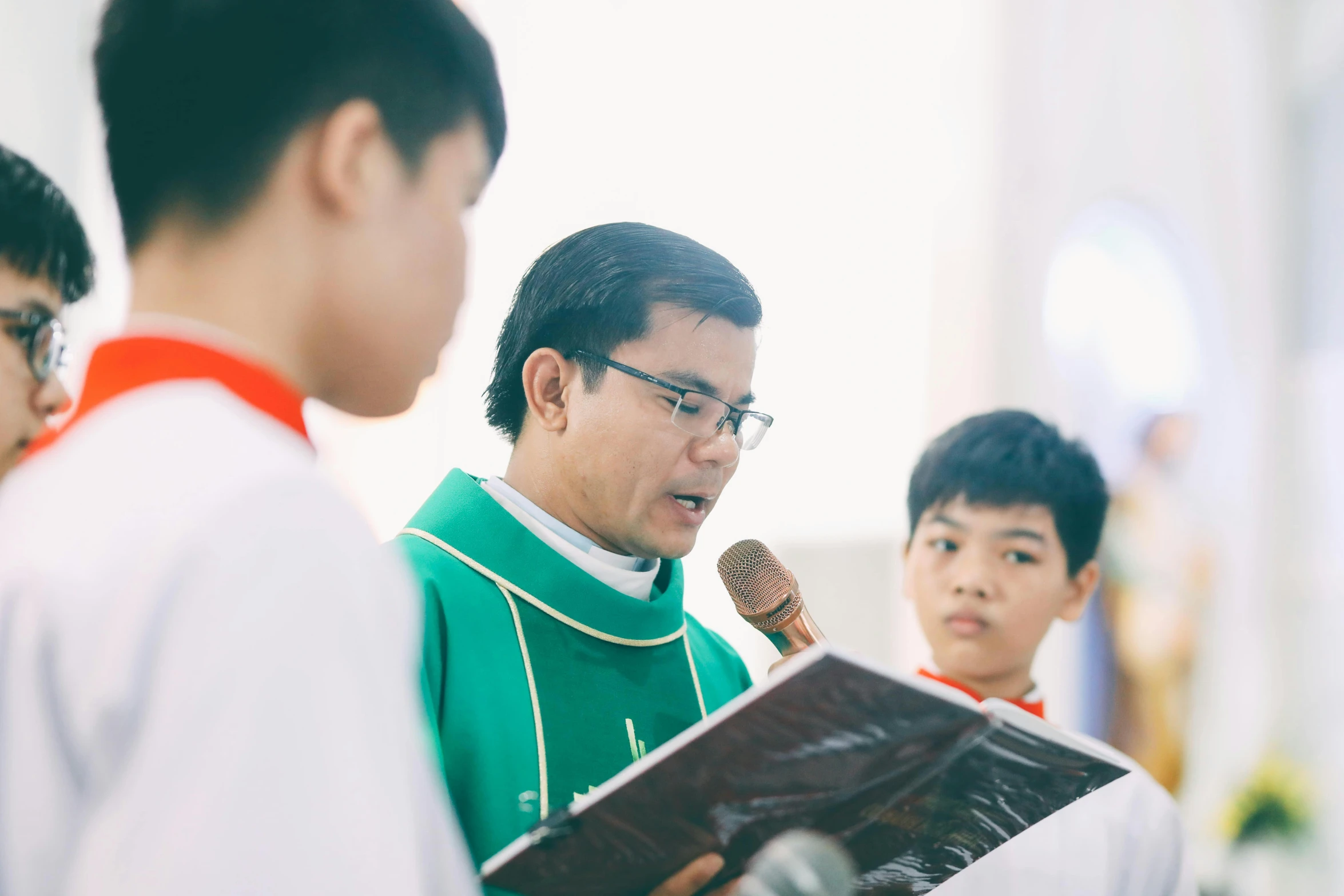 an asian man wearing a green and white clergy outfit holding a microphone in front of him as another boy in red shirt looks at it