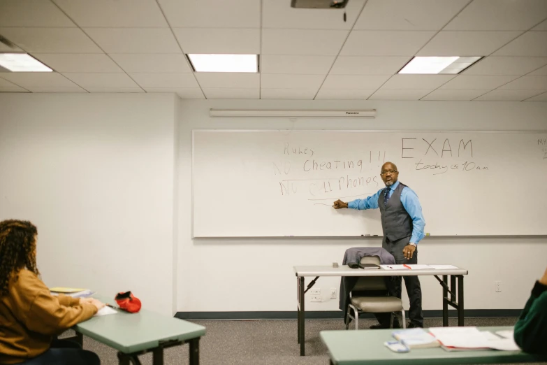 two students are sitting at desks while one teacher stands in front of them