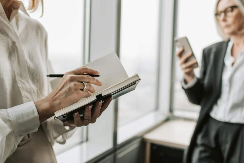 the two women in the office look at the camera with one holding an open notebook and pen