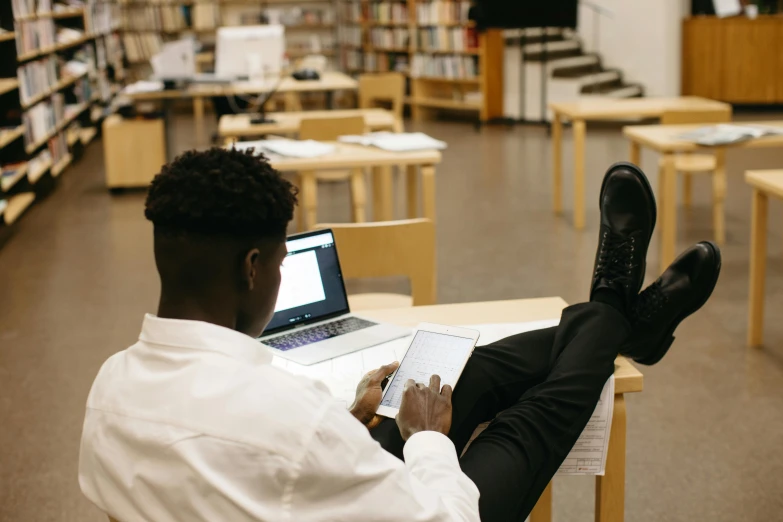 a man in business attire sits in a liry with a laptop and is reading a book