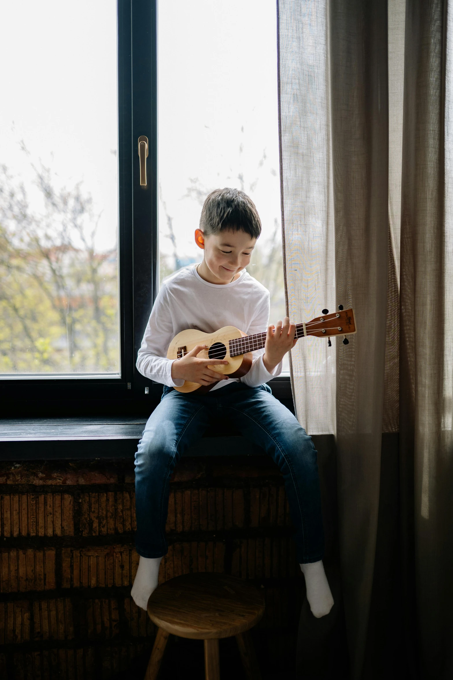 a  sitting next to a window while playing an acoustic guitar