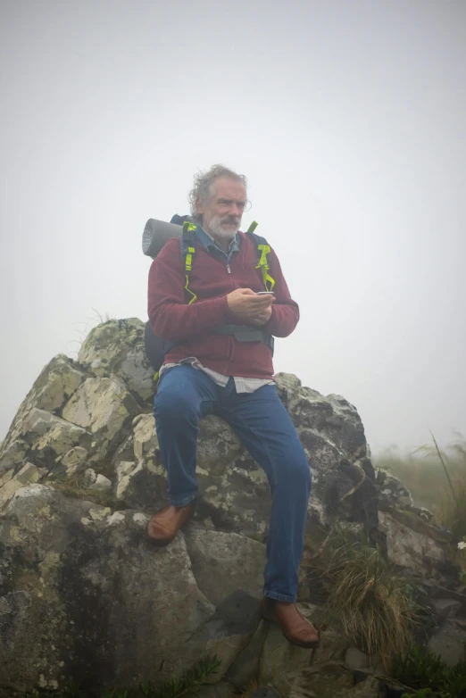 man with backpack sitting on rocks in the fog using cell phone