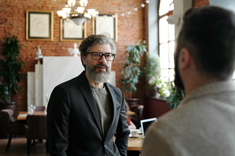 man with beard, glasses and brown sweater speaking to a male in white shirt